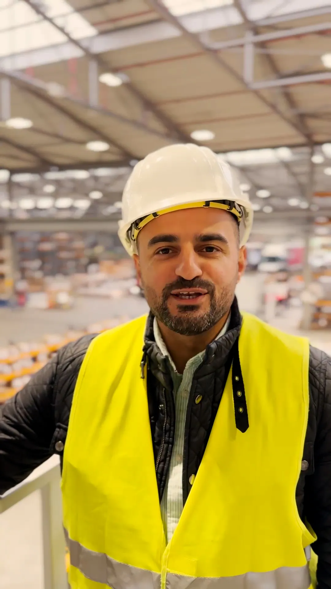Mann mit weißem Schutzhelm und gelber Warnweste blickt in einer hell beleuchteten Industriehalle in die Kamera.