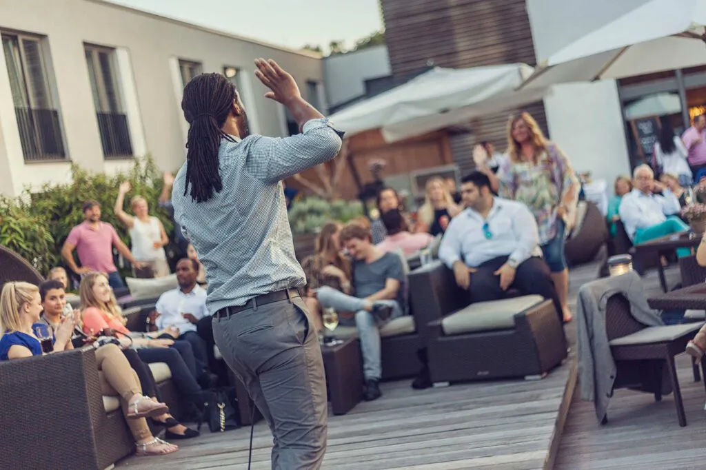 Mann mit Dreadlocks hebt die Hand vor sitzendem Publikum auf der sonnigen Terrasse des Aspria Uhlenhorst.