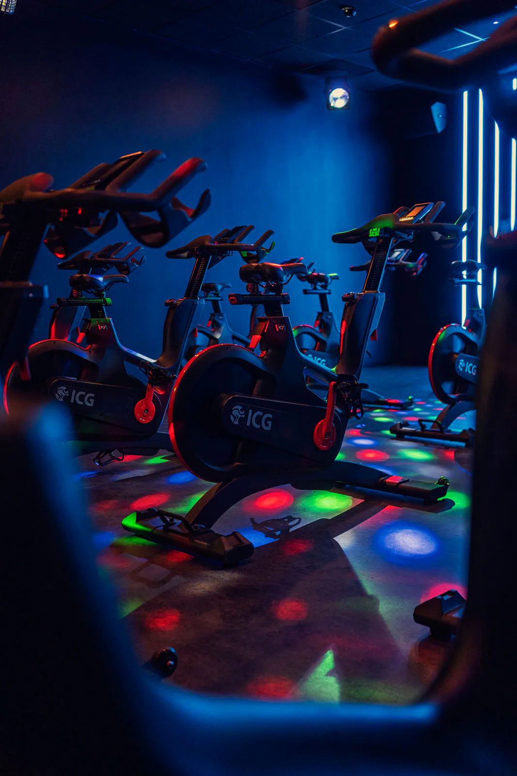 Spinningräder in einem dunklen Fitnessstudio mit blauem Licht und bunten Lichtpunkten auf dem Boden.