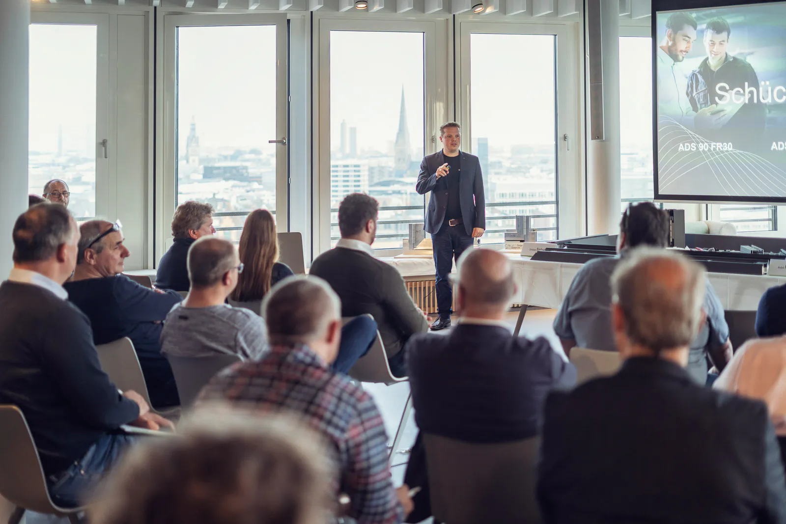 Redner mit Mikrofon vor sitzendem Publikum bei Schüco-Event in hellem Raum mit großen Fenstern und Präsentationsleinwand.