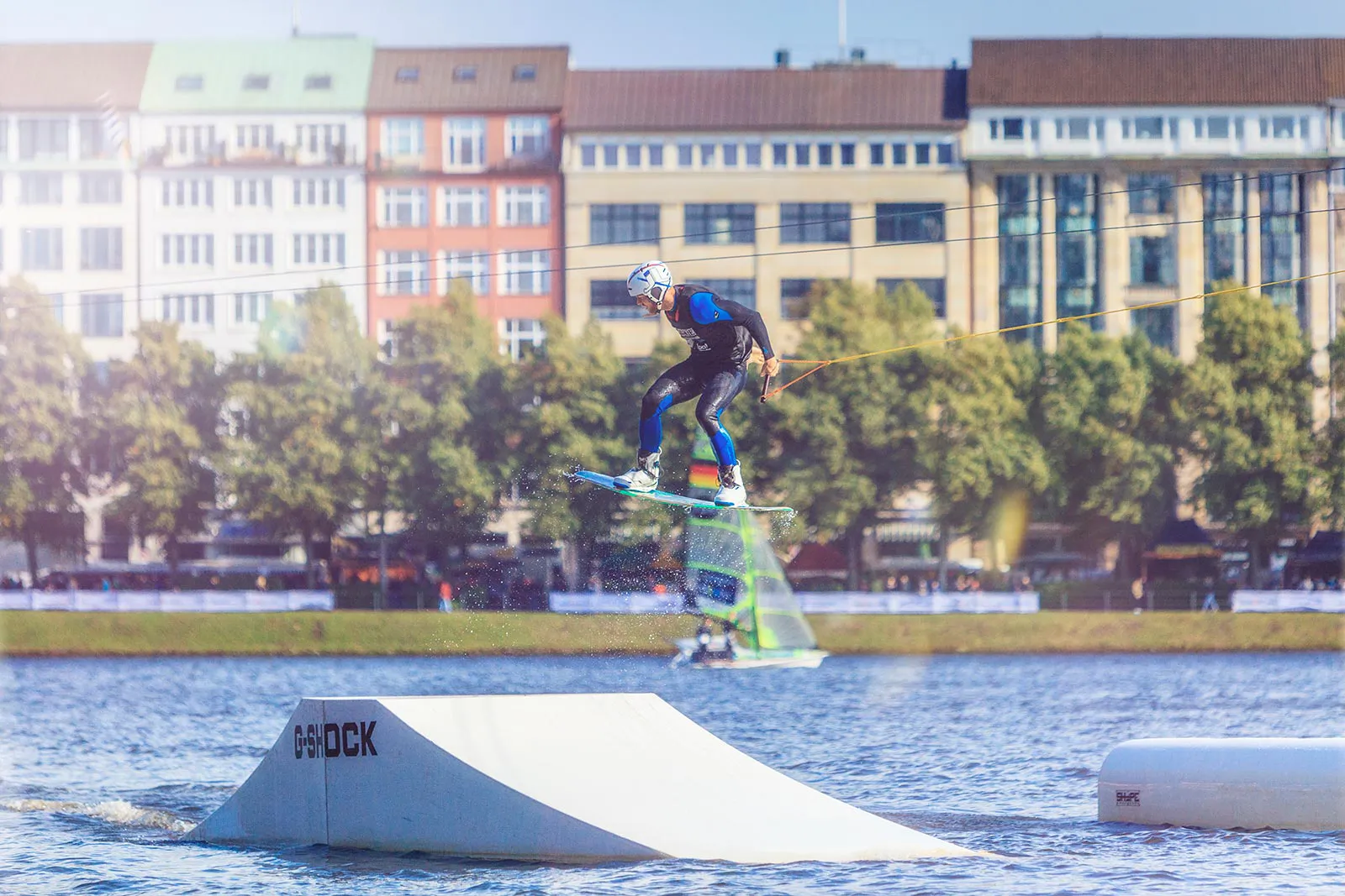 Wakeboarder springt bei Sonnenlicht über eine G-Shock-Rampe auf dem Wasser, vor Bäumen und Häusern.