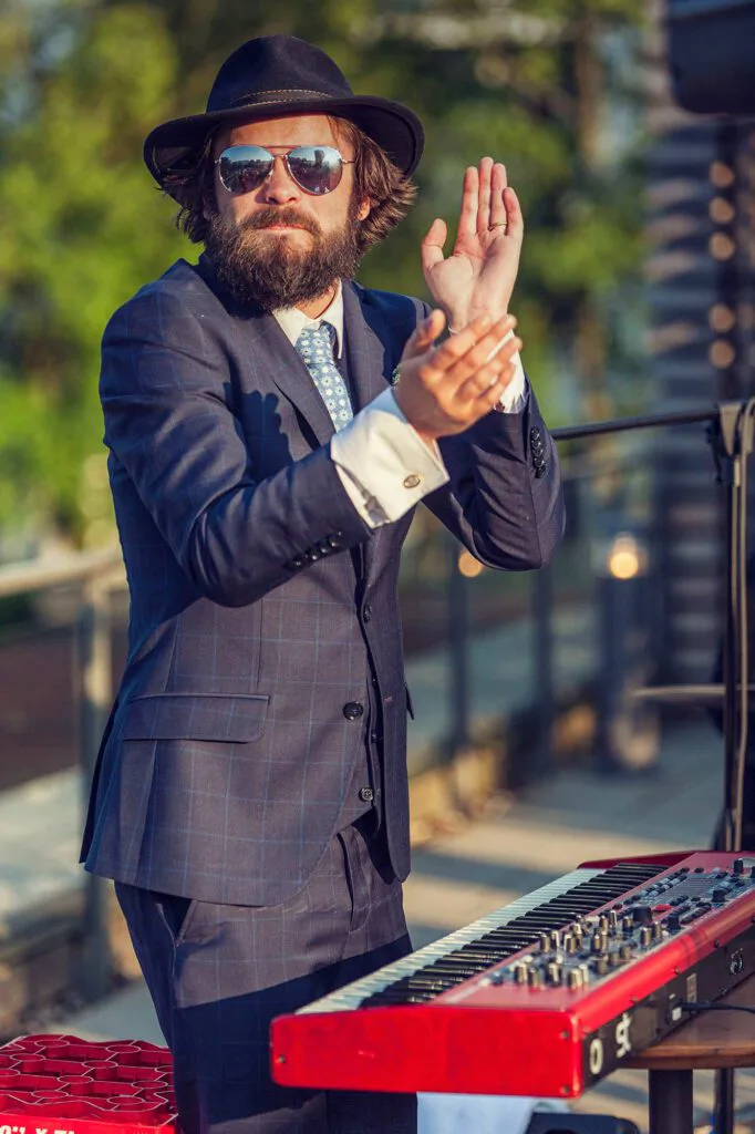 Bärtiger Musiker mit Hut und Sonnenbrille klatscht im Sonnenlicht hinter einem roten Keyboard im Freien.