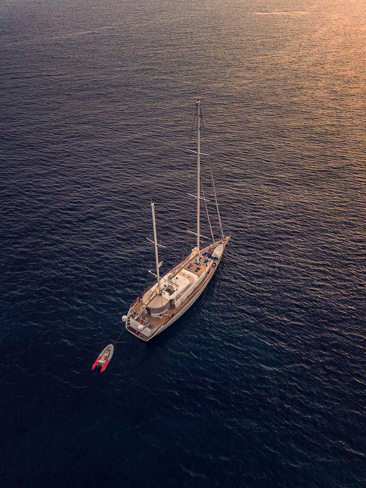 Luftaufnahme einer Segelyacht mit kleinem Beiboot auf dunklem Wasser im warmen Abendlicht.