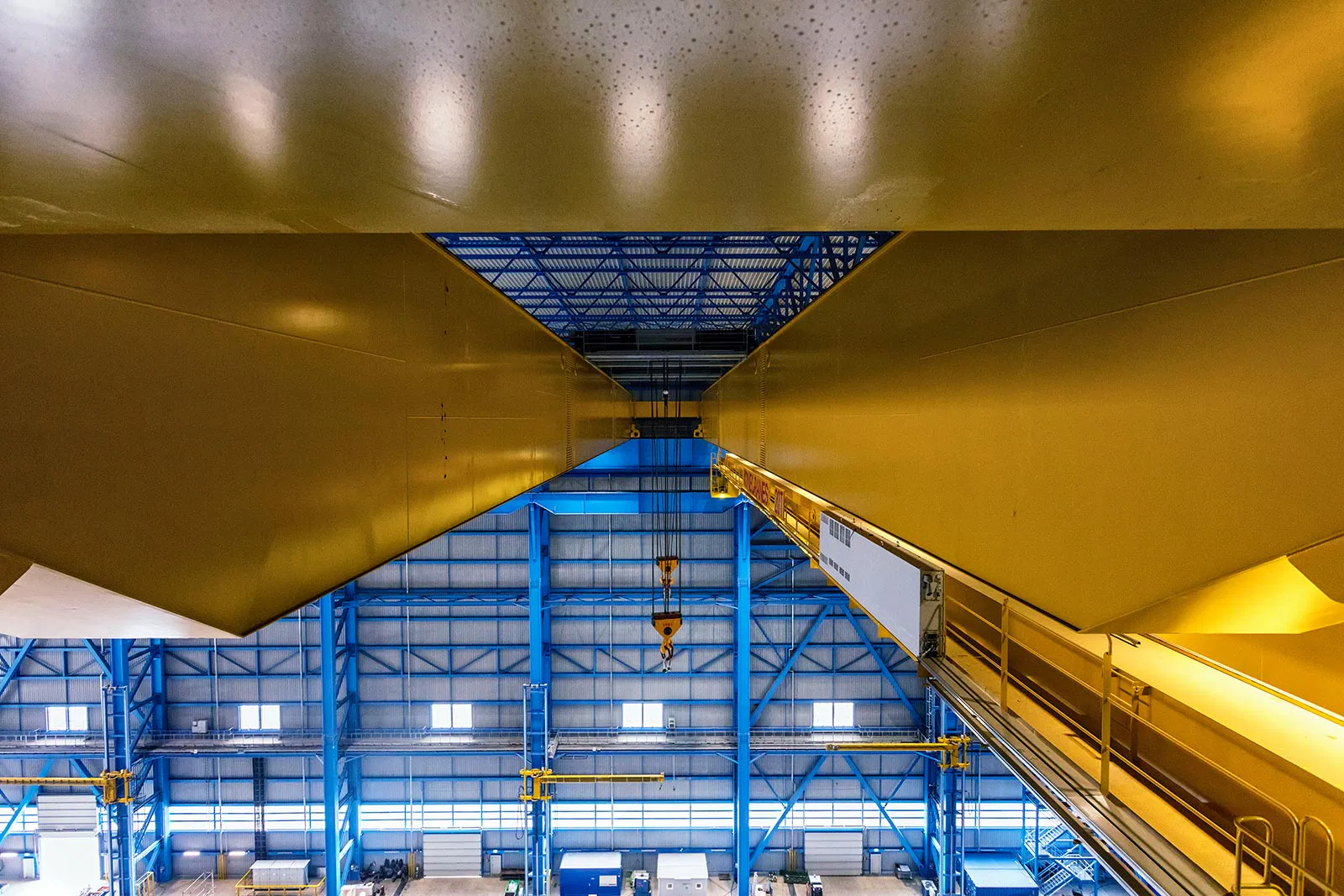 Gelbe Kranträger mit Laufsteg und hängendem Haken in einer tageslichthellen Industriehalle mit blauen Stahlträgern.