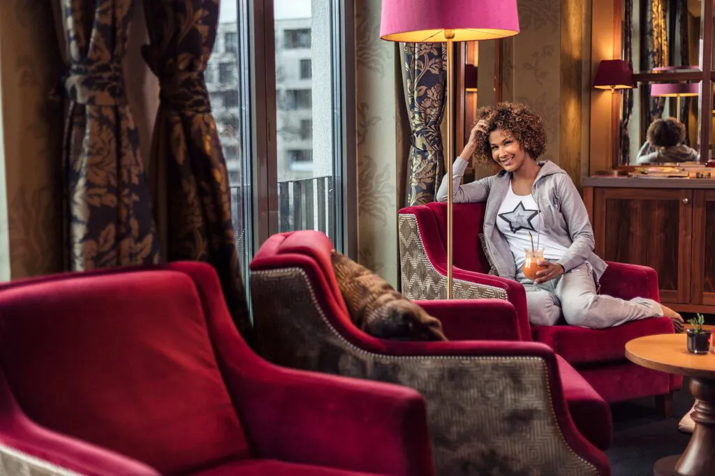 Marie Amière sitzt mit einem Getränk im roten Sessel einer Lounge, neben einem Fenster und warm leuchtenden Lampen.