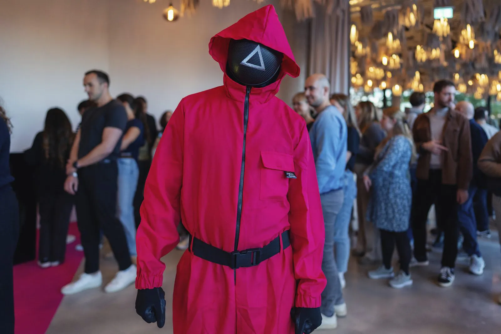 Person im pinken Kapuzenanzug mit schwarzer Dreiecksmaske vor Gästen unter warmen Pendelleuchten beim Netflix-Event.