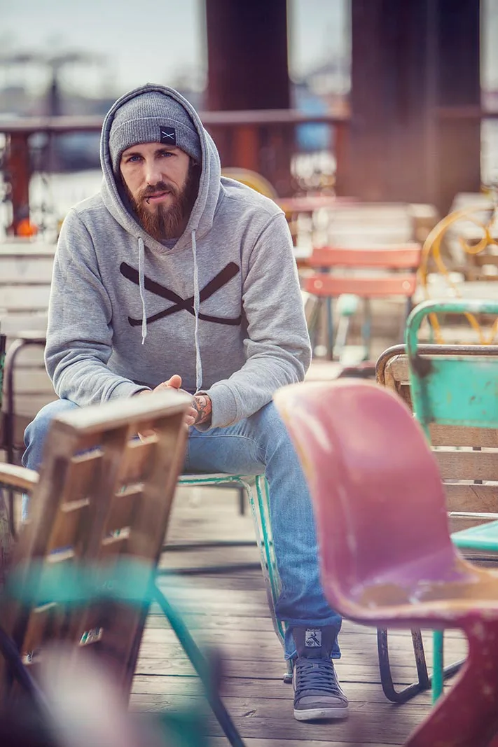 Bärtiger Mann mit Mütze und grauem Hoodie sitzt bei Tageslicht zwischen bunten Stühlen auf einer Holzterrasse.