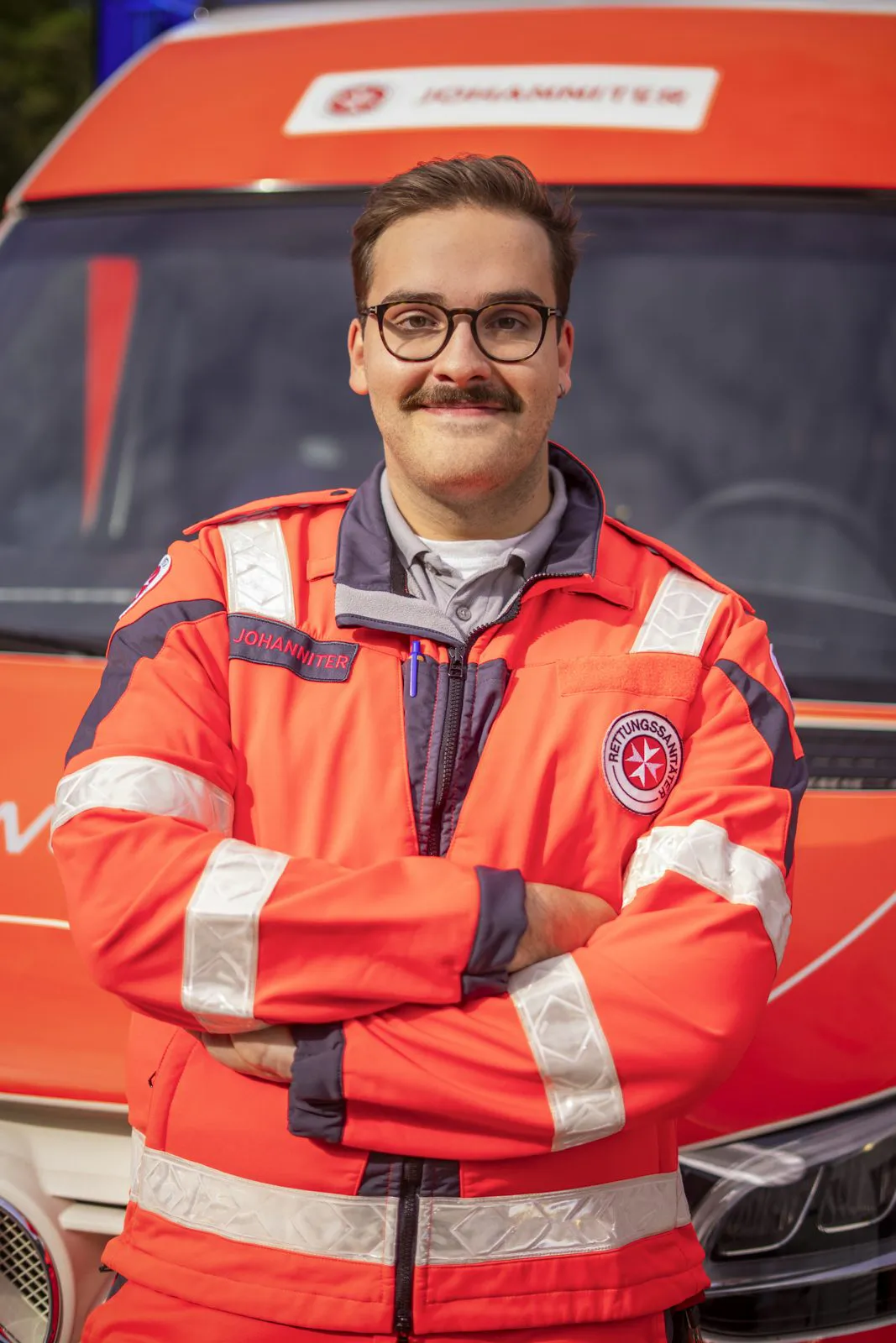 Johanniter-Rettungskraft mit Brille und Schnurrbart steht mit verschränkten Armen vor einem Rettungswagen.