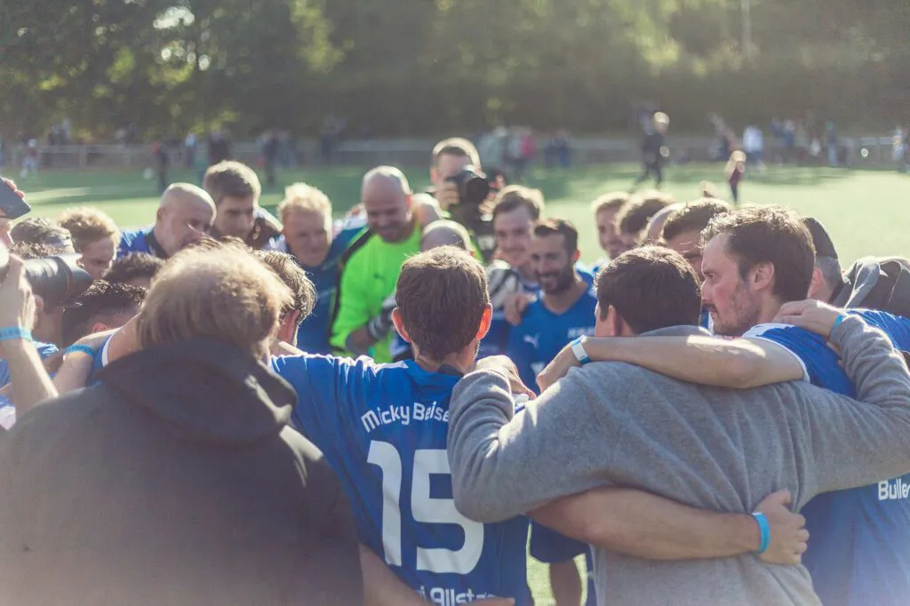 Fußballspieler in blauen Trikots stehen Arm in Arm im Mannschaftskreis auf einem sonnigen Sportplatz.