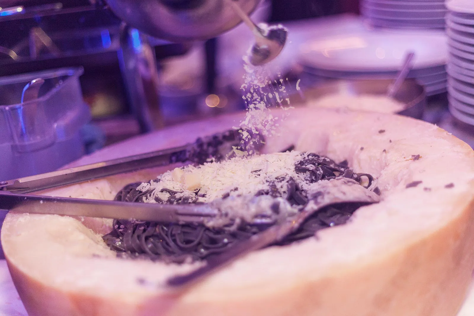 Dunkle Pasta in einem ausgehöhlten Käselaib wird am Buffet bei violettem Licht mit geriebenem Käse bestreut.