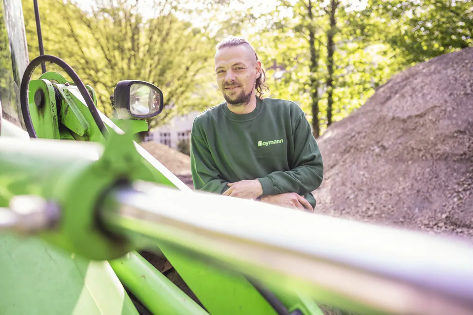 Mann im grünen Boymann-Pullover lehnt bei Sonnenlicht an einer grünen Baumaschine, dahinter Schotter und Bäume.
