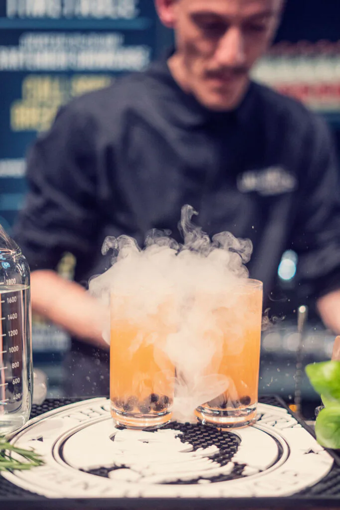 Barkeeper hinter zwei orangefarbenen Drinks mit aufsteigendem weißem Nebel am Thomas-Henry-Stand beim OMR Festival.