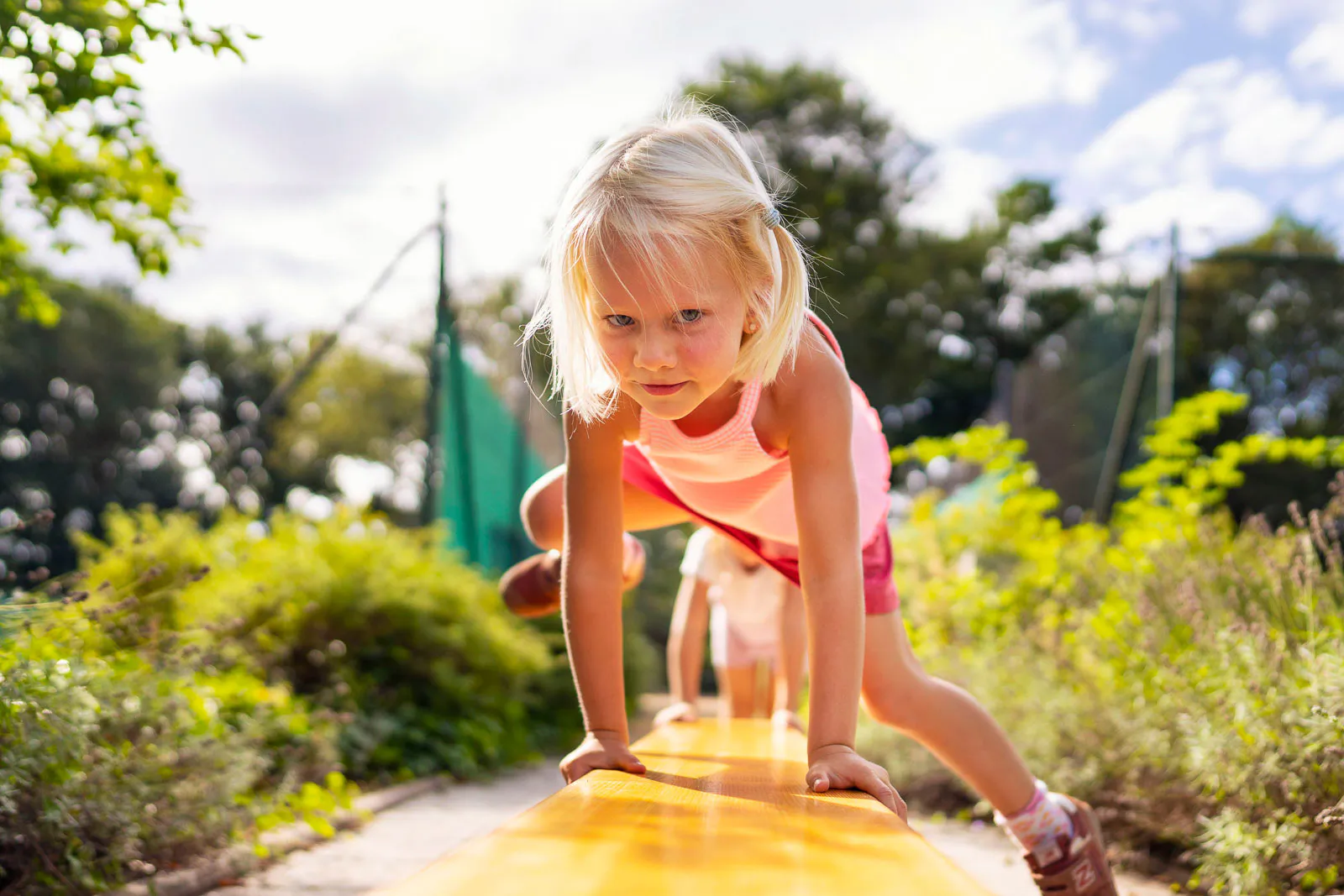 Blondes Kind klettert im Sonnenlicht über einen gelben Balancierbalken auf einem begrünten Spielplatz.