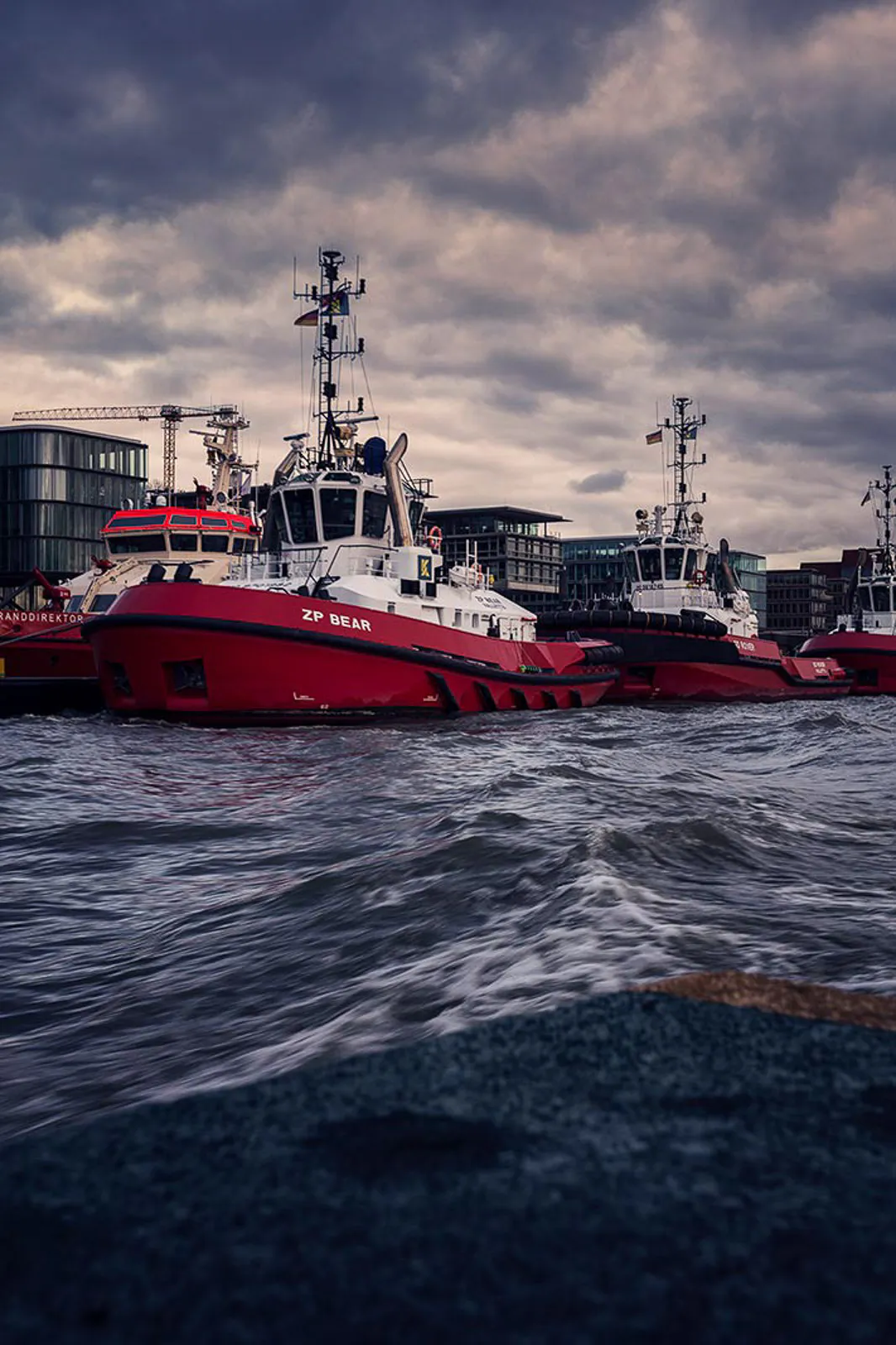 Rote Schlepper liegen vor Hafengebäuden unter dunklen Wolken, im Vordergrund welliges Wasser.