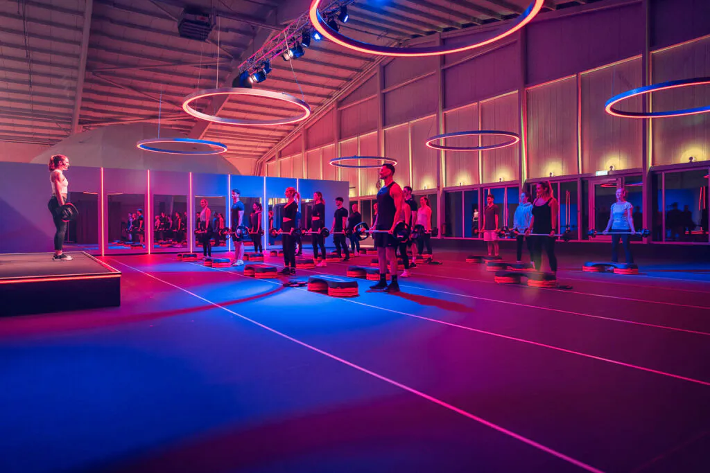 Gruppenkurs an Stepbrettern in einer großen Trainingshalle unter mehreren rot und blau leuchtenden Lichtringen.