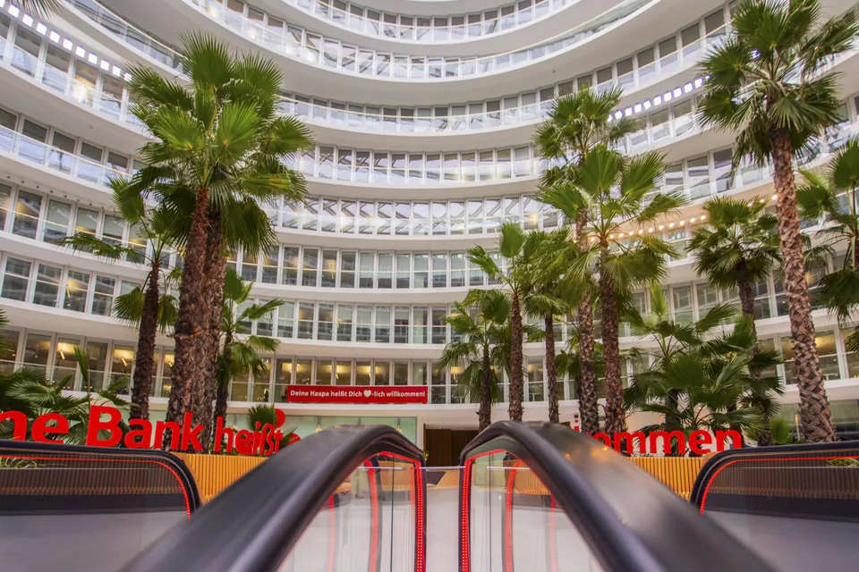 Rolltreppen führen in ein helles Atrium mit hohen Palmen und geschwungenen, mehrstöckigen Fensterfronten.