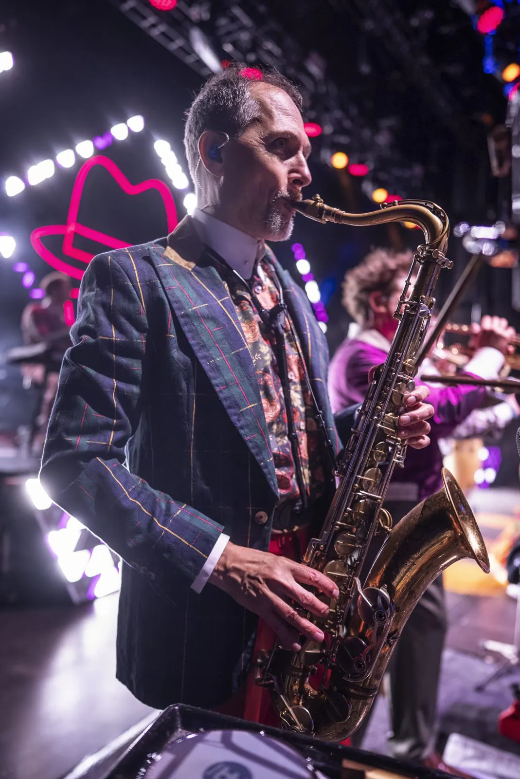 Saxofonist im karierten Sakko spielt auf einer bunt beleuchteten Konzertbühne, dahinter weitere Musiker.