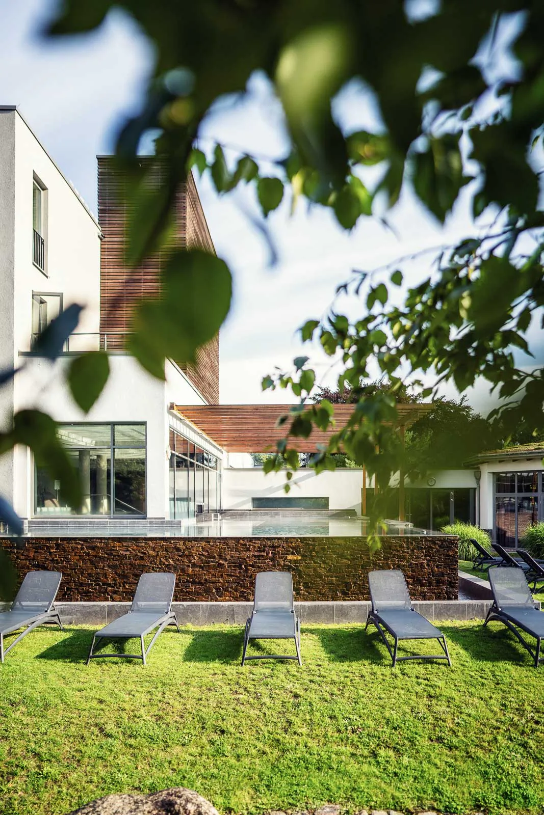 Sonnenliegen auf grünem Rasen vor modernem Saunagebäude mit Wasserbecken, eingerahmt von Blättern im Sonnenlicht.
