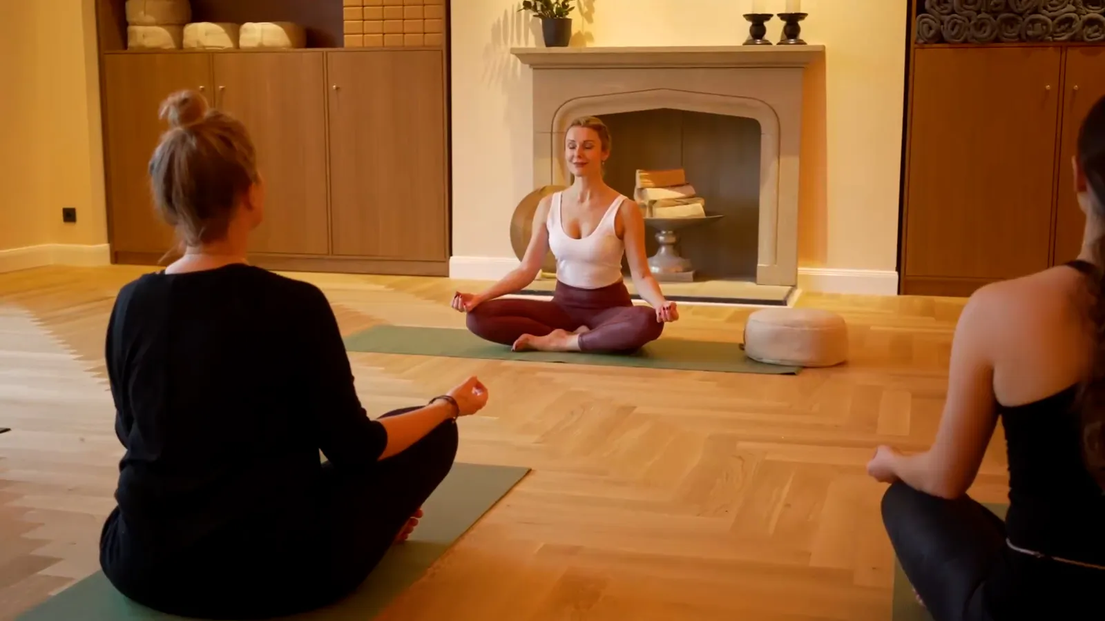 Drei Frauen sitzen im Schneidersitz auf Yogamatten in einem warm beleuchteten Raum mit Parkett und Kamin.
