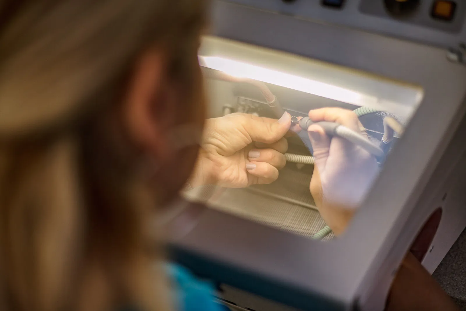Person bearbeitet ein kleines Werkstück mit einem Handgerät an einem beleuchteten Arbeitsplatz im Dentallabor.